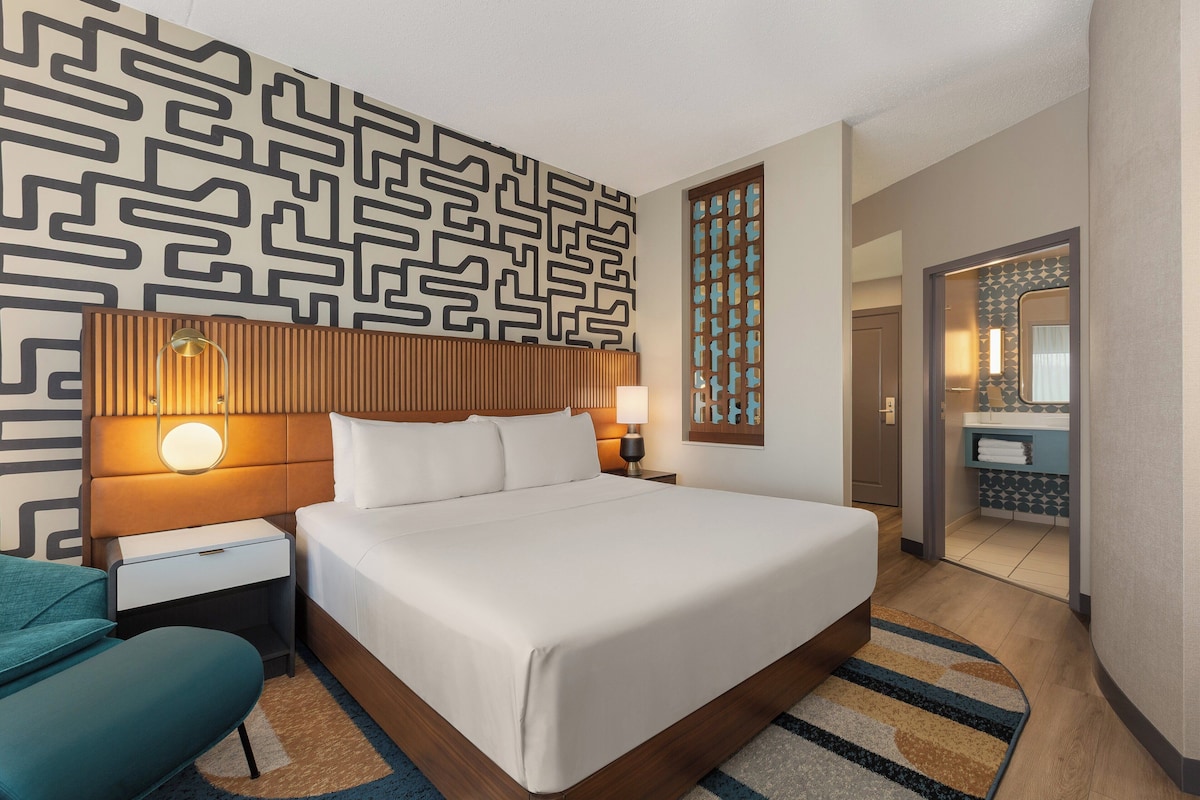 A hotel room with a large bed, a nightstand with a lamp, a wooden headboard, and a patterned wall.