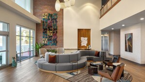 A spacious lobby with a curved gray sectional sofa, a small round table, and a seating area with leather chairs. A large window provides natural light, and a colorful abstract wall art is visible. - Hotel Indigo Columbus Architectural Center by IHG (Columbus)