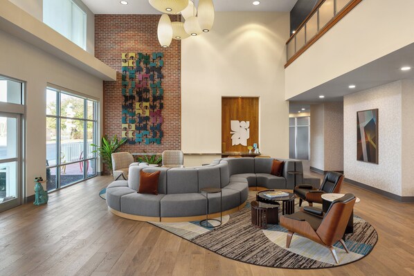 A spacious lobby with a curved gray sectional sofa, a small round table, and a seating area with leather chairs. A large window provides natural light, and a colorful abstract wall art is visible. - Hotel Indigo Columbus Architectural Center by IHG (Columbus)