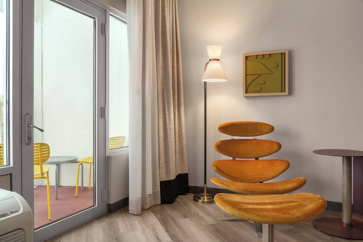 A modern room with a sliding glass door leading to an outdoor area, a floor lamp, a unique orange chair, and a minimalist wall art piece.