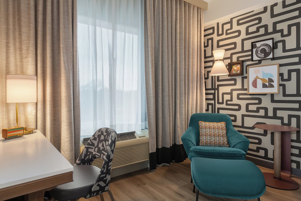 A hotel room with a modern design, featuring a teal armchair, a white desk, a standing lamp, and a patterned wall.