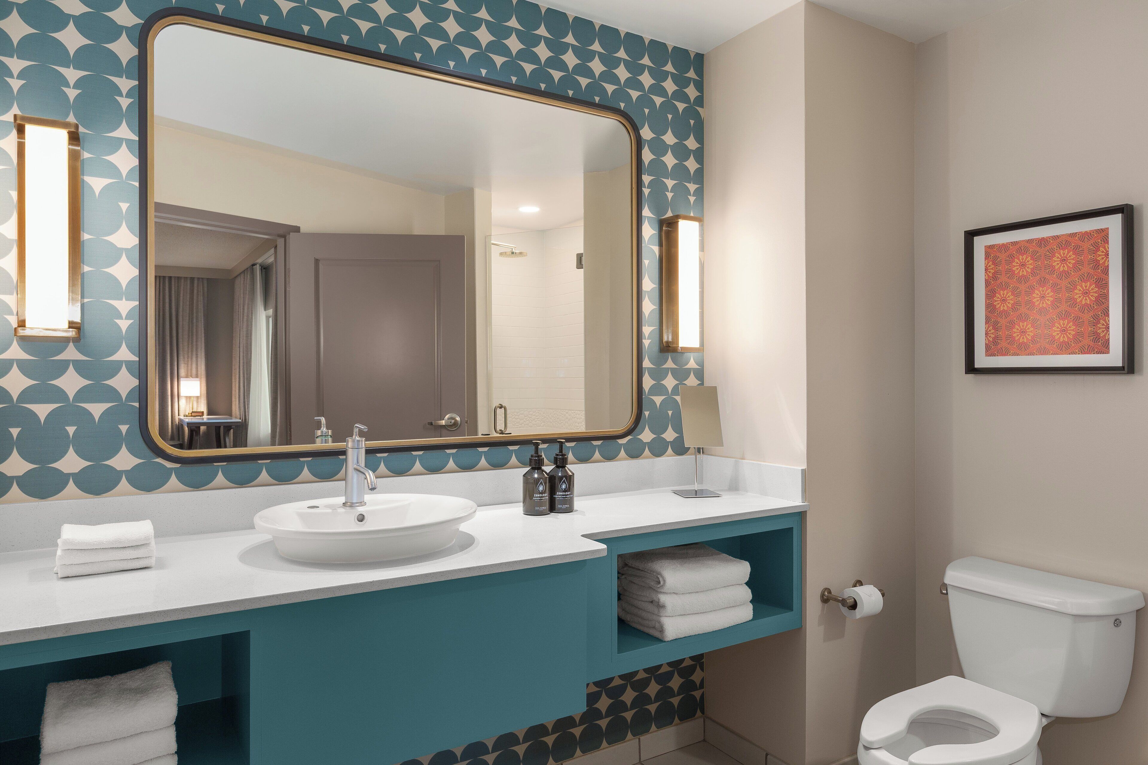 A bathroom with a large mirror, white sink, teal vanity, white toilet, and a framed picture on the wall.