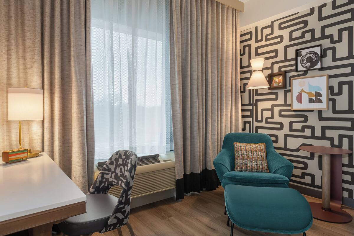 A hotel room with a modern design, featuring a teal armchair, a white desk, a standing lamp, and a patterned wall.