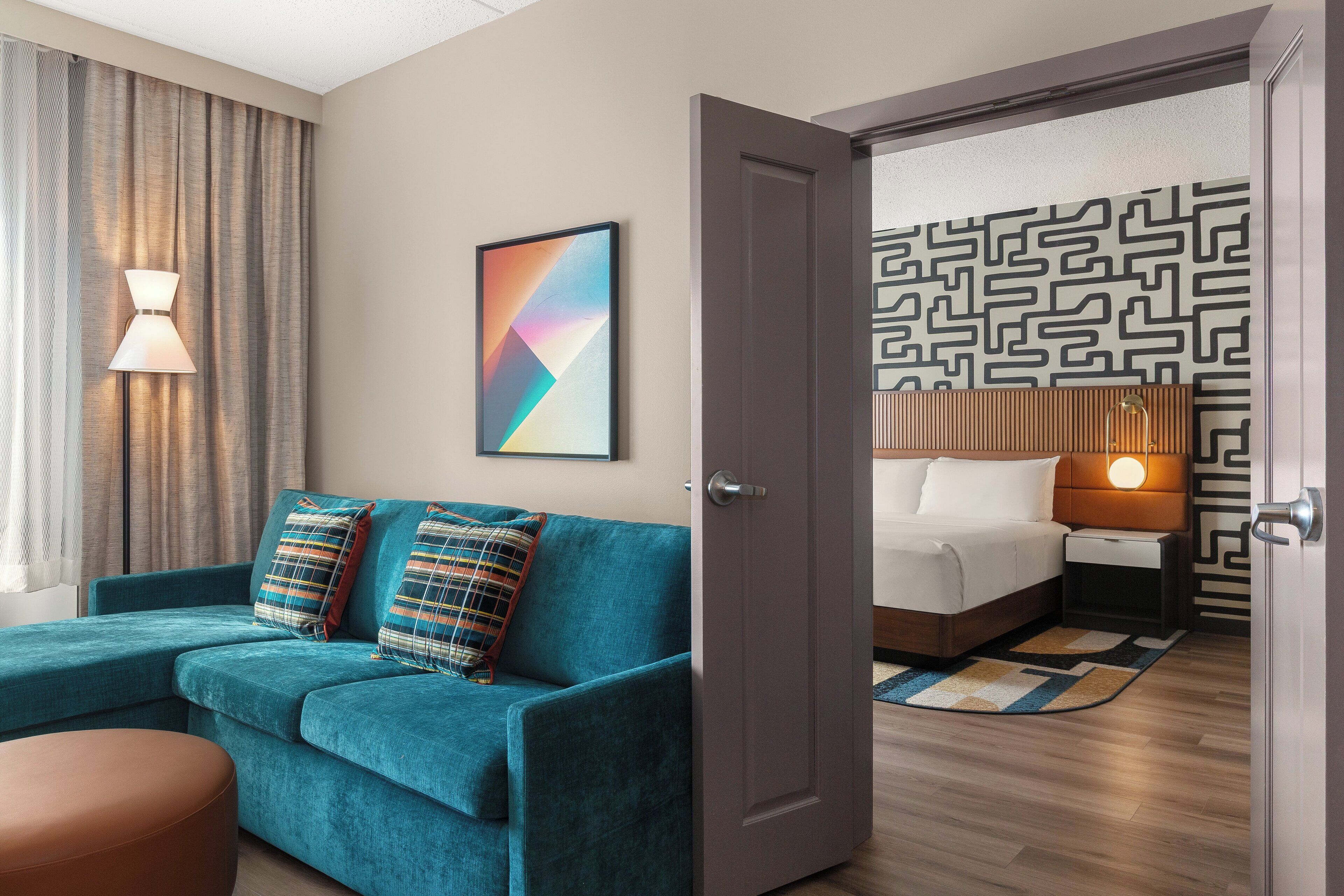 A modern hotel room with a teal sofa, a bed with a wooden headboard, a patterned wall, and a door leading to another room.