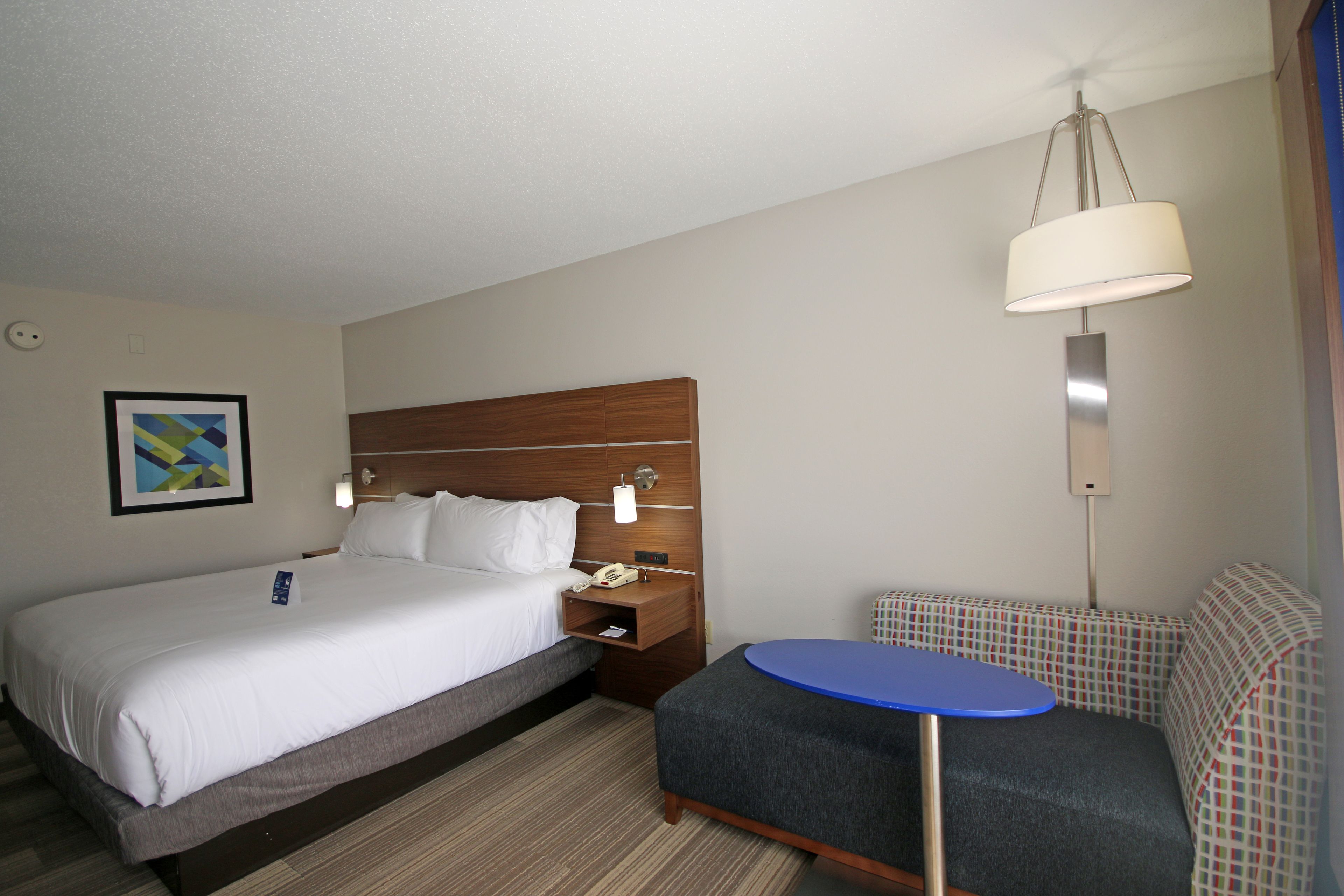 A hotel room with a large bed, a bedside table with a lamp, a small round table, and a patterned bench.