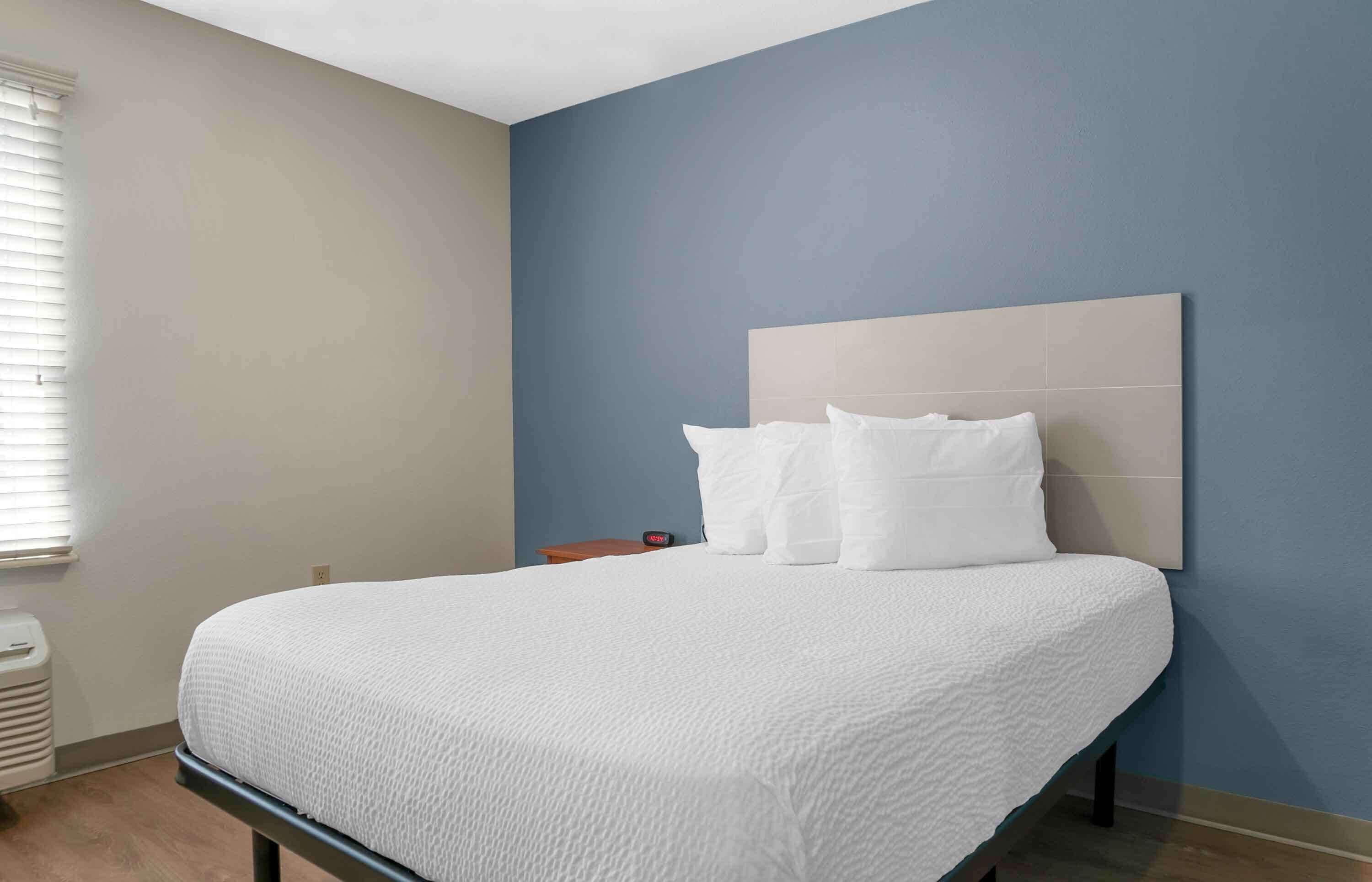 Una cama bien tendida con un edredón blanco y almohadas, una mesita de noche de madera con un reloj, y una pared azul.