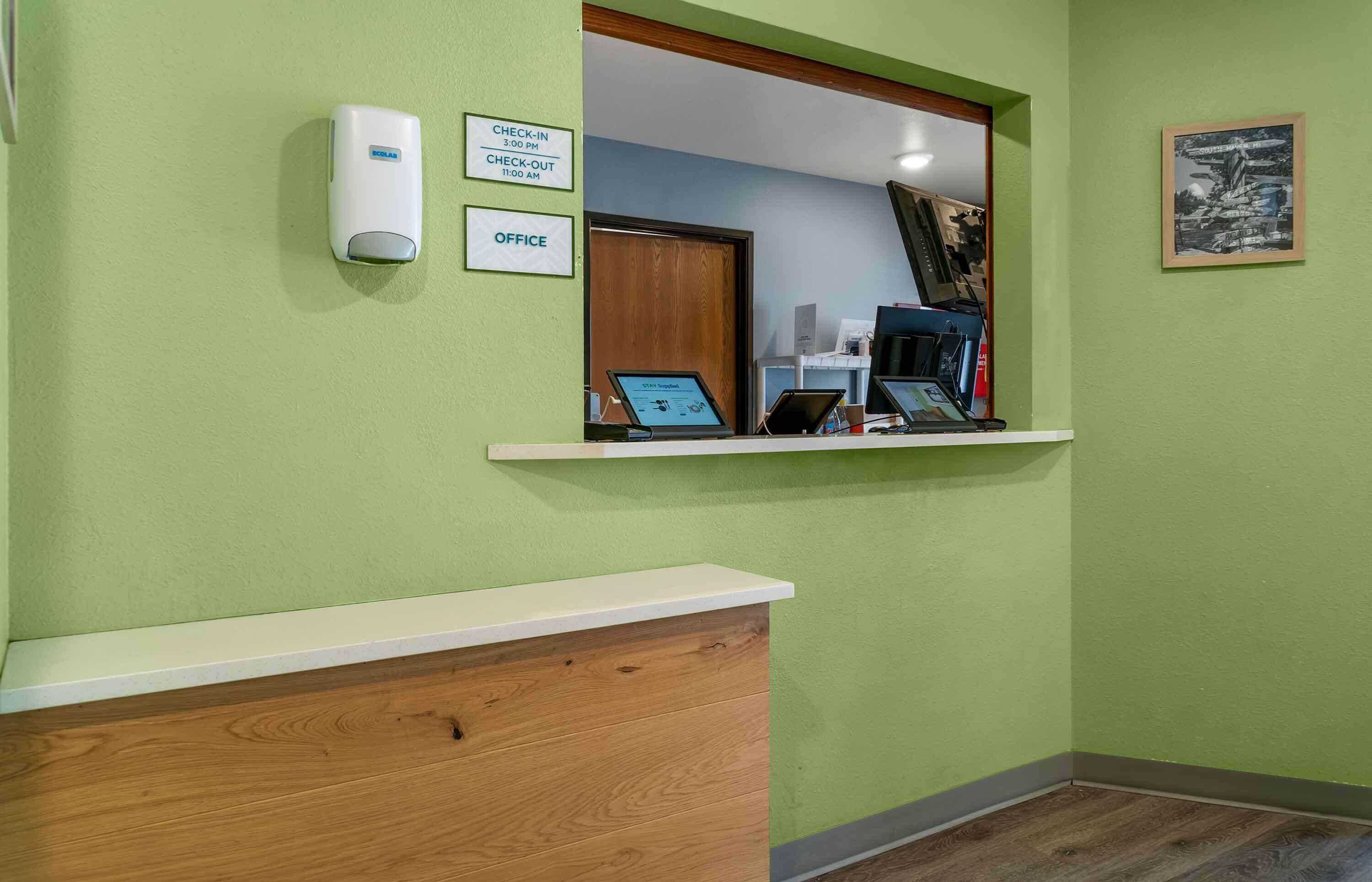 Un mostrador de recepción con un cartel de registro de entrada y salida, un dispensador de jabón y un cuadro en la pared.