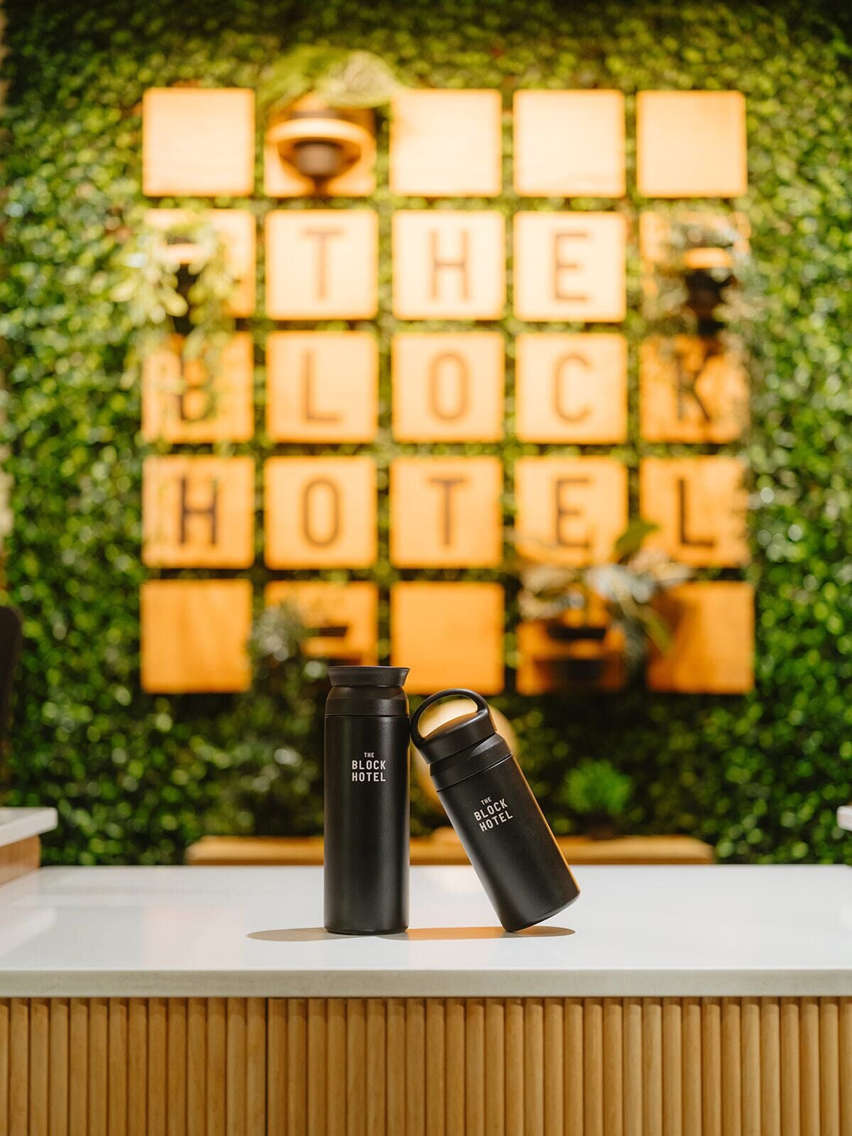 A reception desk with two black water bottles labeled 'The Block Hotel' and a wall with illuminated letters spelling 'THE BLOCK HOTEL'.