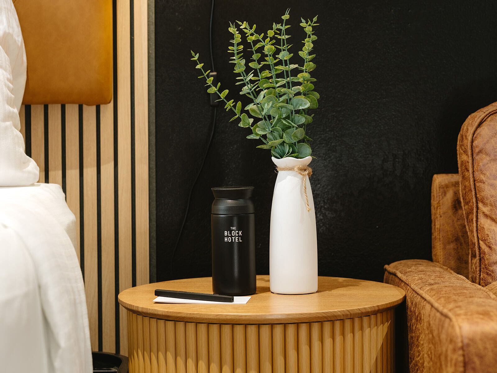 Een rond houten bijzettafeltje met een zwart tumblerglas, een witte vaas met een plant, en een boek.