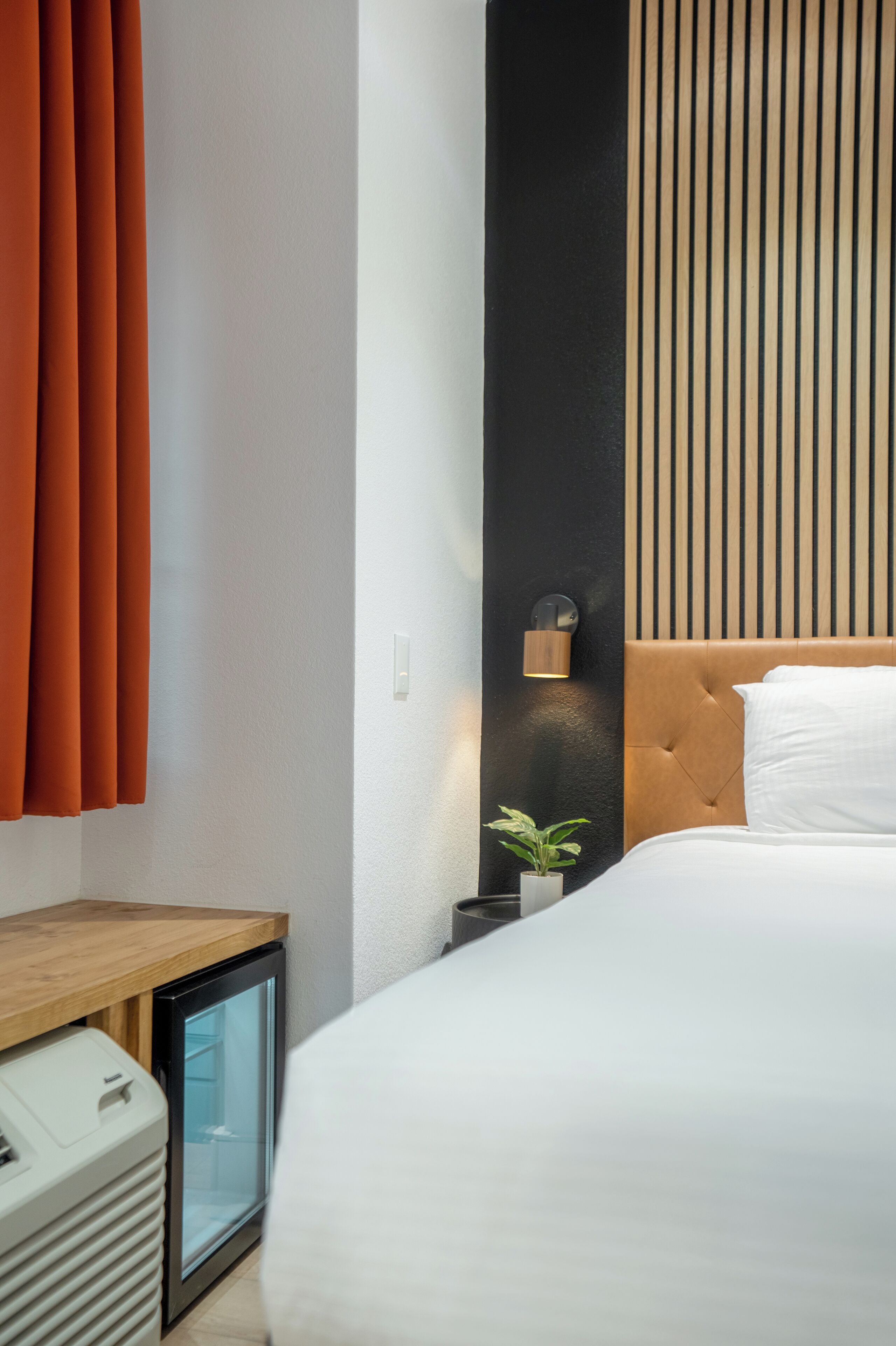 A modern hotel room with a bed, a small bedside table, a nightstand, a wall-mounted lamp, and a potted plant.