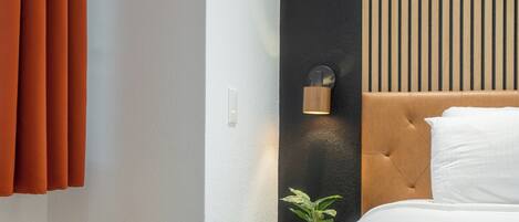 A modern hotel room with a bed, a small bedside table, a nightstand, a wall-mounted lamp, and a potted plant.