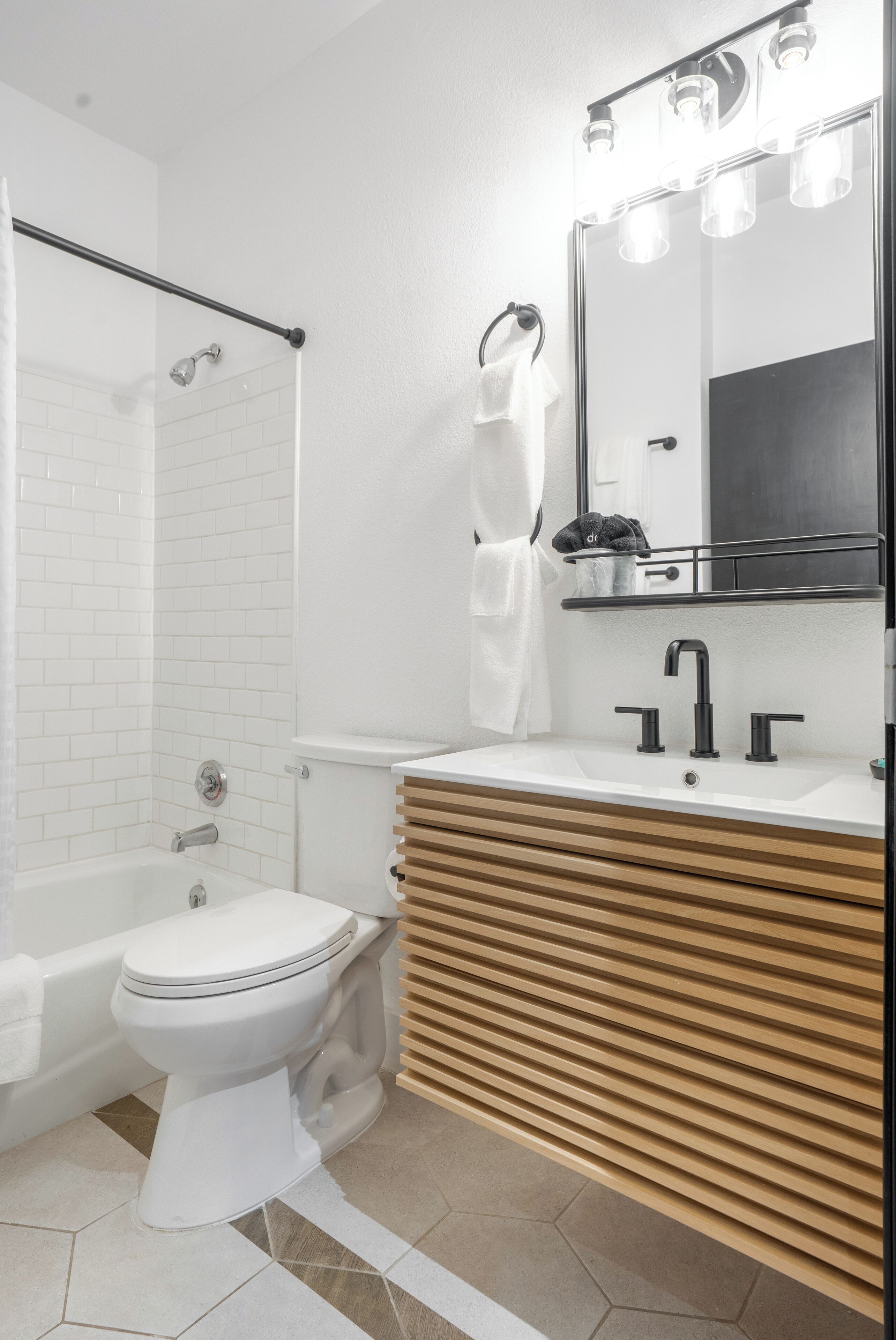 A modern bathroom with a white toilet, a wooden vanity, a large mirror with lights, a shower area, and a towel rack.