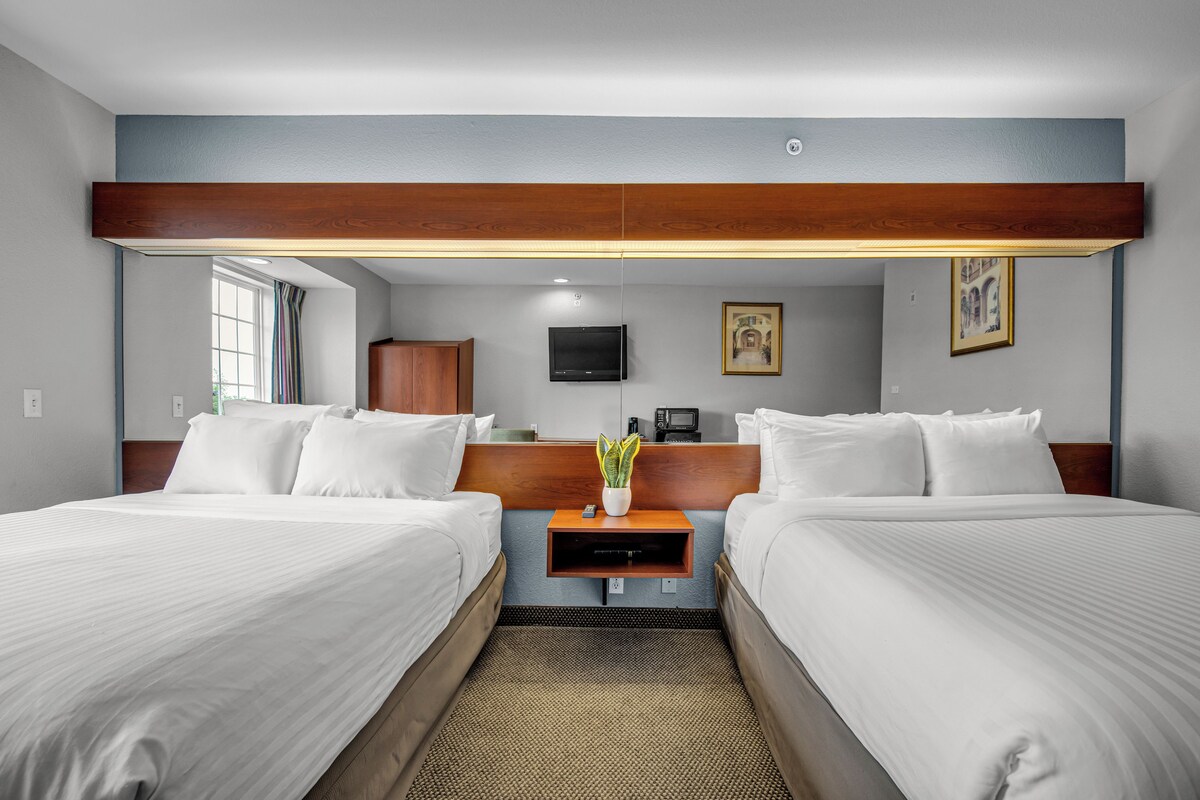 A hotel room with two beds, a wooden headboard, a flat-screen TV, a small table with a plant, and a window with curtains.