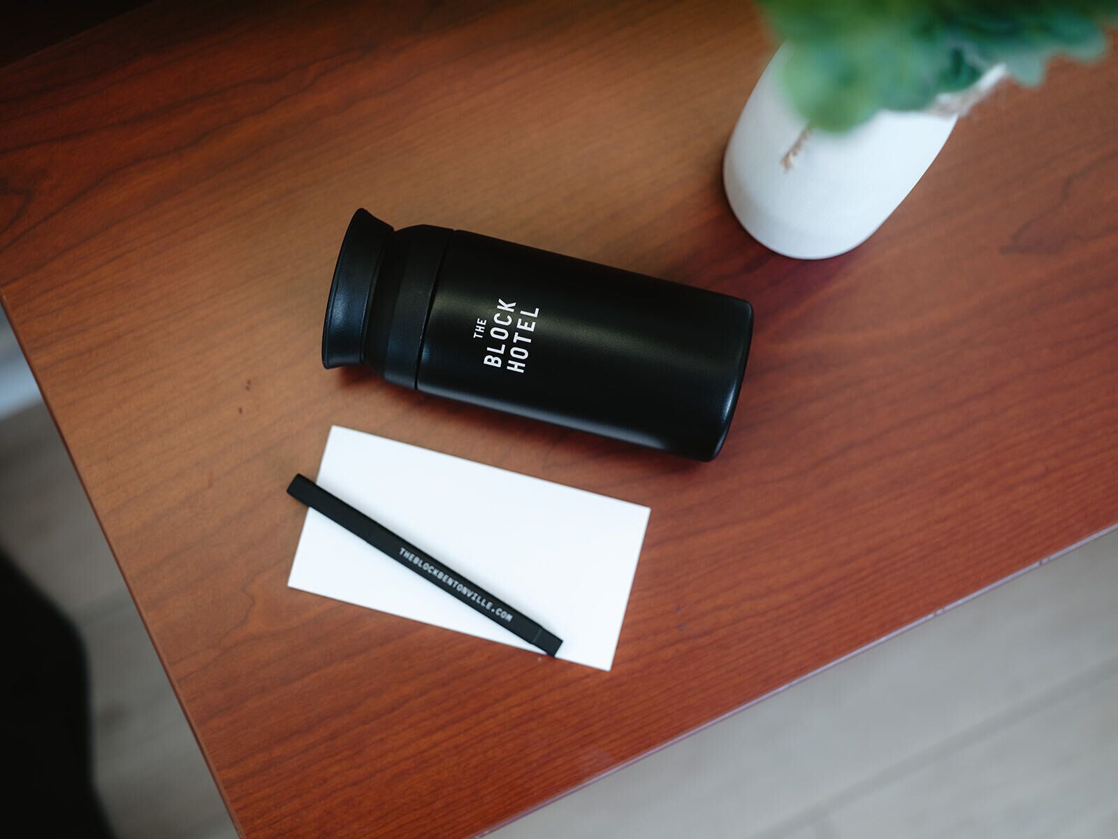 Een zwarte waterfles met de tekst 'The Block Hotel' op een houten oppervlak, met een wit kaartje en een pen ernaast.