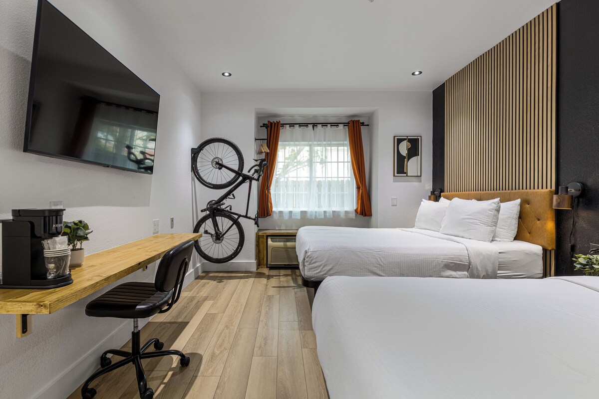 Une chambre d’hôtel avec un bureau en bois, un vélo, deux lits, une télévision et une fenêtre avec des rideaux.