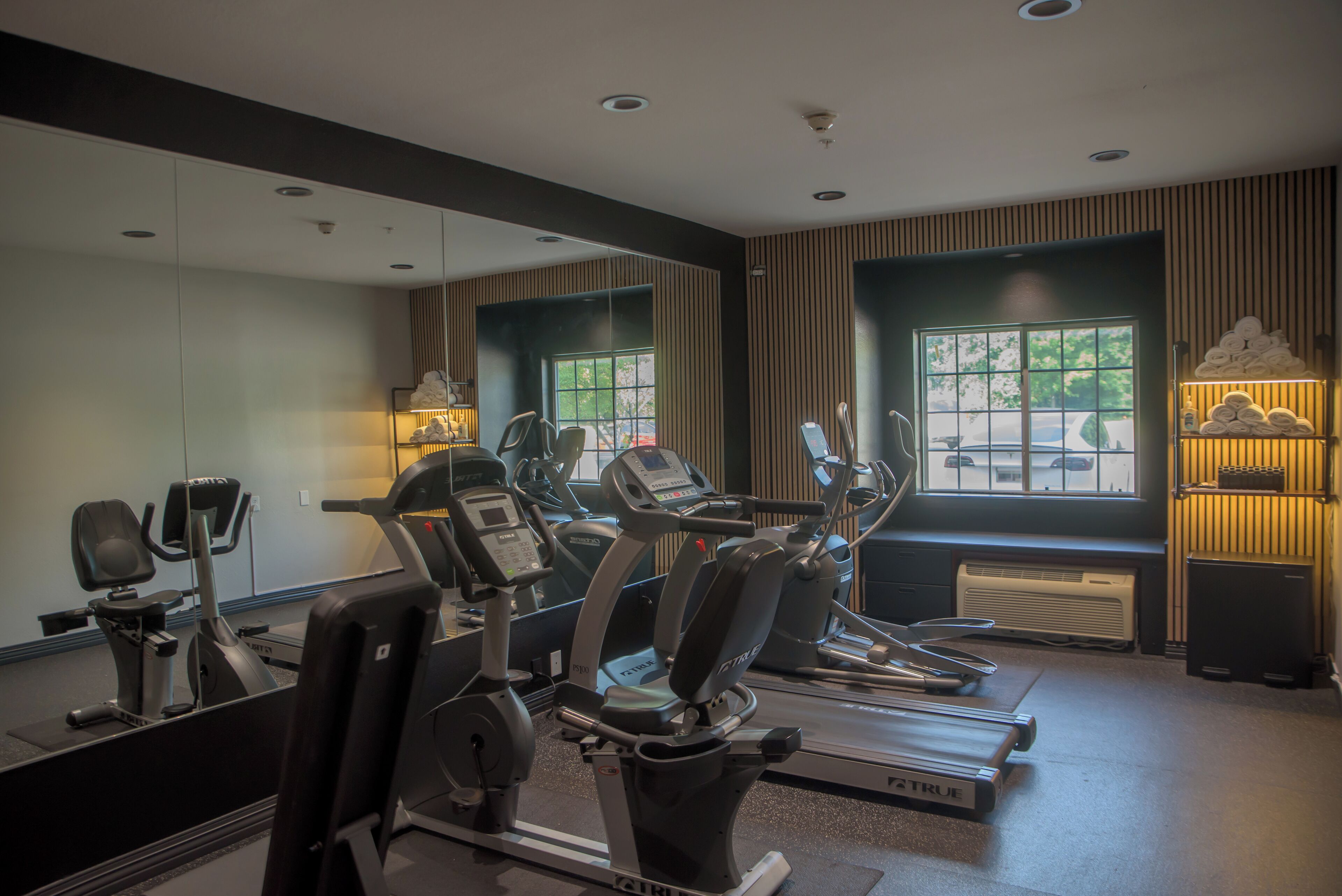 Une salle de sport équipée de machines de cardio, dotée d’un mur avec des miroirs et d’une fenêtre donnant sur les voitures garées.
