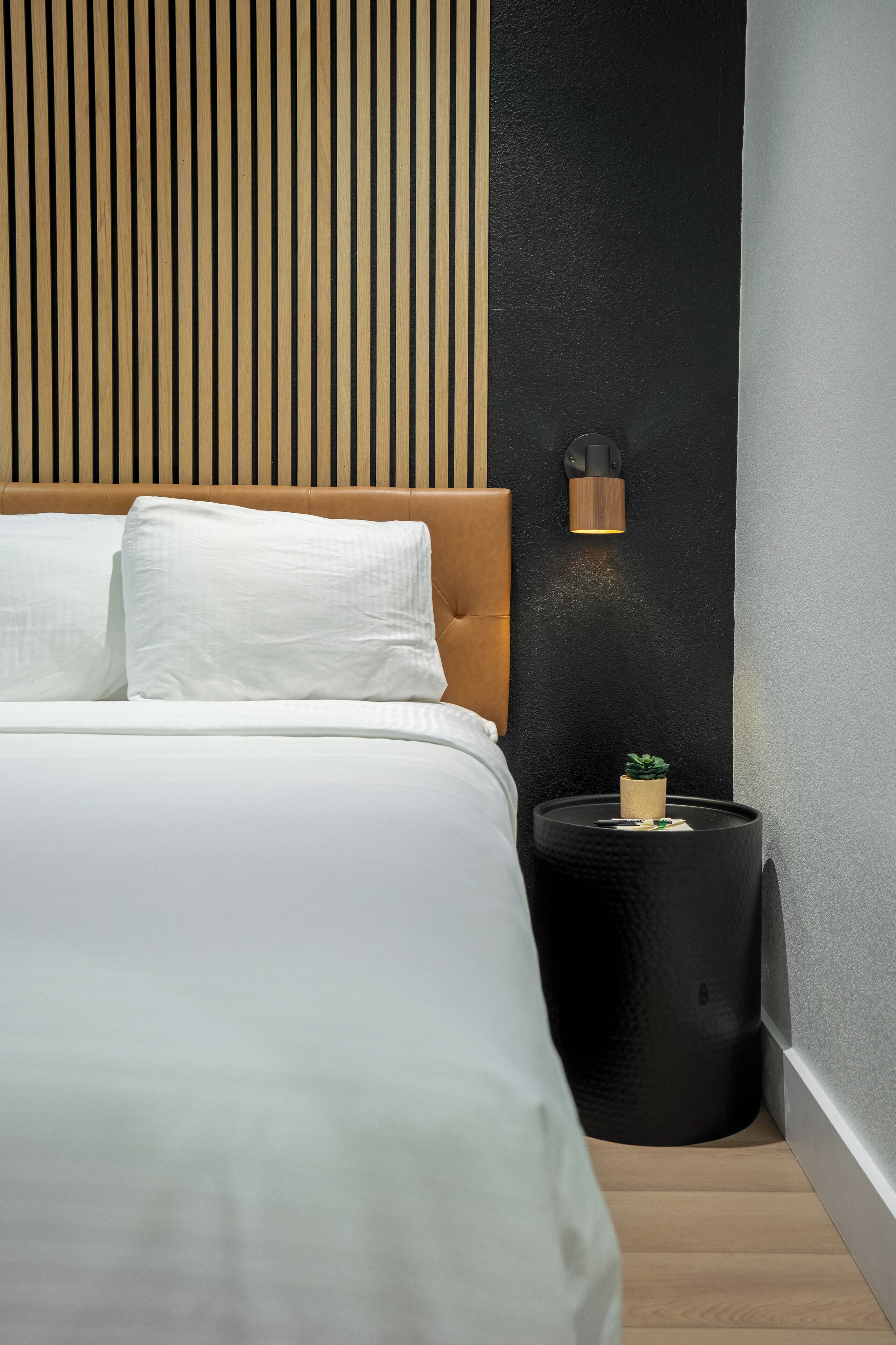 Un dormitorio moderno con cabecera de madera, ropa de cama blanca, mesita de noche negra con textura y una pequeña planta en maceta.