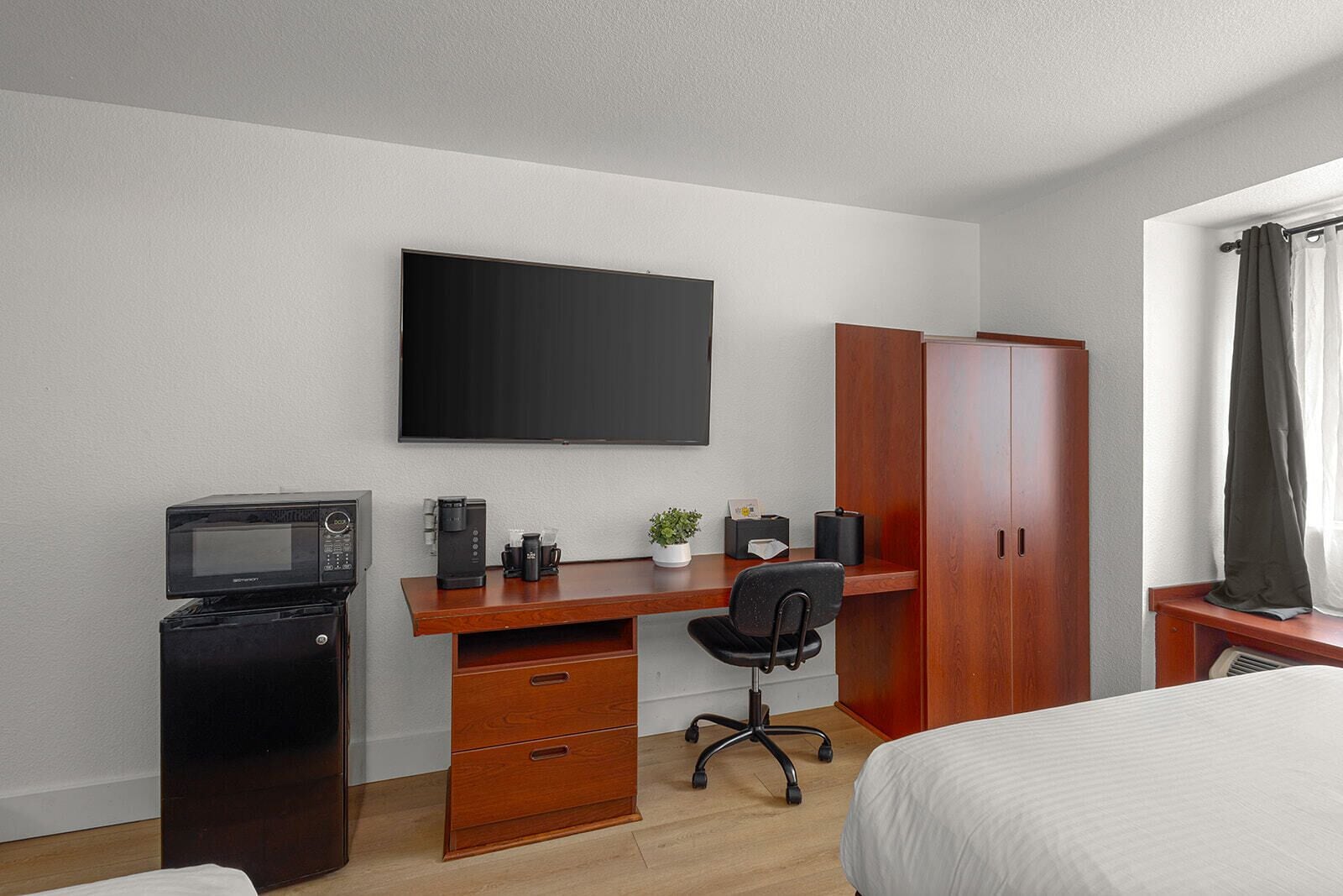A hotel room with a wooden desk, a black office chair, a flat-screen TV, a microwave, a small potted plant, a wardrobe, and a bed.