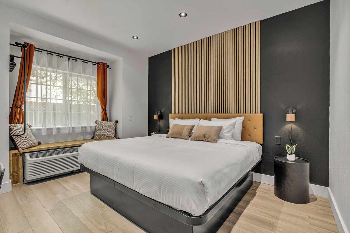 A modern bedroom with a large bed, a bench by the window, and a wall-mounted lamp.
