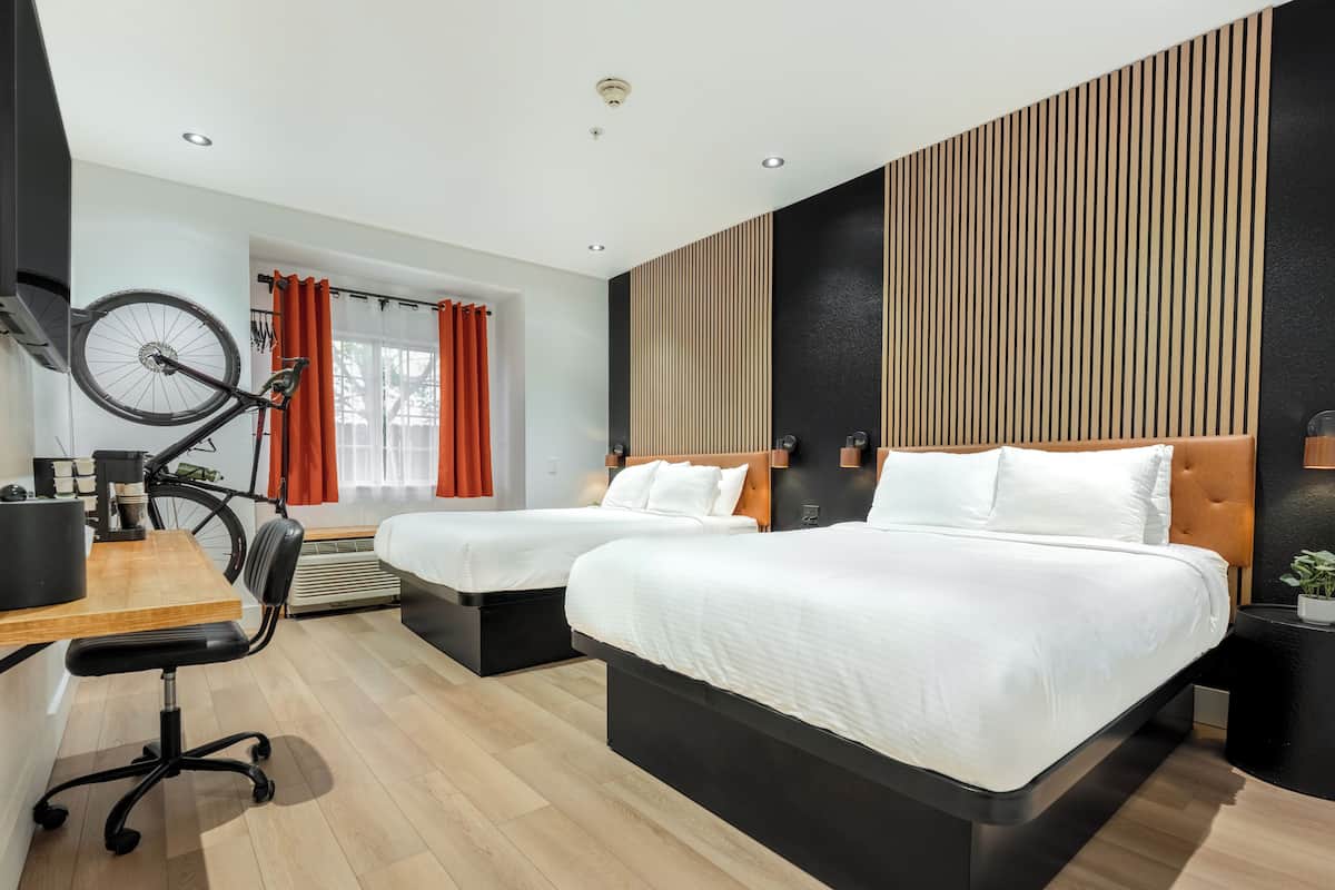 A modern hotel room with two beds, a desk with a chair, and a bicycle.