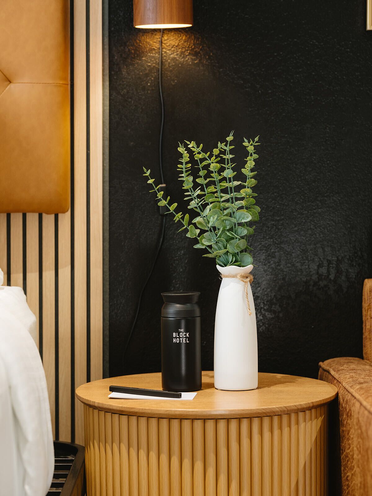 Une table ronde en bois sur laquelle se trouvent un vase blanc contenant des feuilles vertes, un thermos noir et une télécommande.