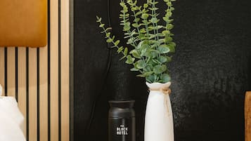 Une table ronde en bois sur laquelle se trouvent un vase blanc contenant des feuilles vertes, un thermos noir et une télécommande.