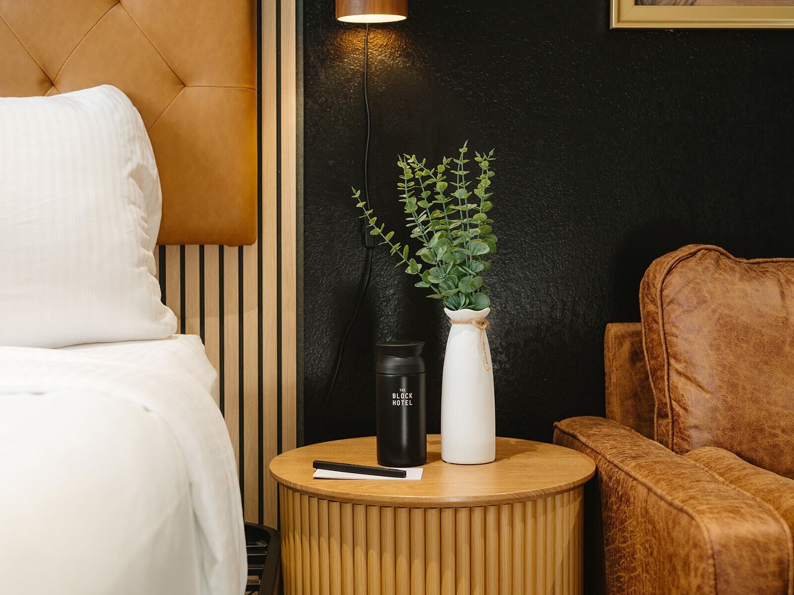 A bed, a round wooden nightstand with a black and white vase, a black tumbler, and a brown leather chair.
