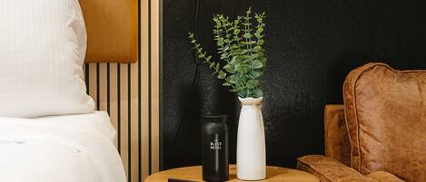 A bed, a round wooden nightstand with a black and white vase, a black tumbler, and a brown leather chair.