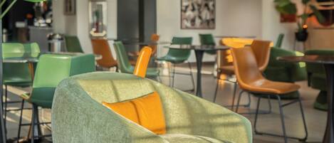 A modern lounge area with green and orange chairs, a round wooden table with a plant, and a large green sofa with an orange cushion.
