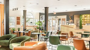 Ein modernes Café mit runden Tischen und grün-orangefarbenen Stühlen, ausgestattet mit großen Fenstern und einer grünen Wand mit Pflanzen.