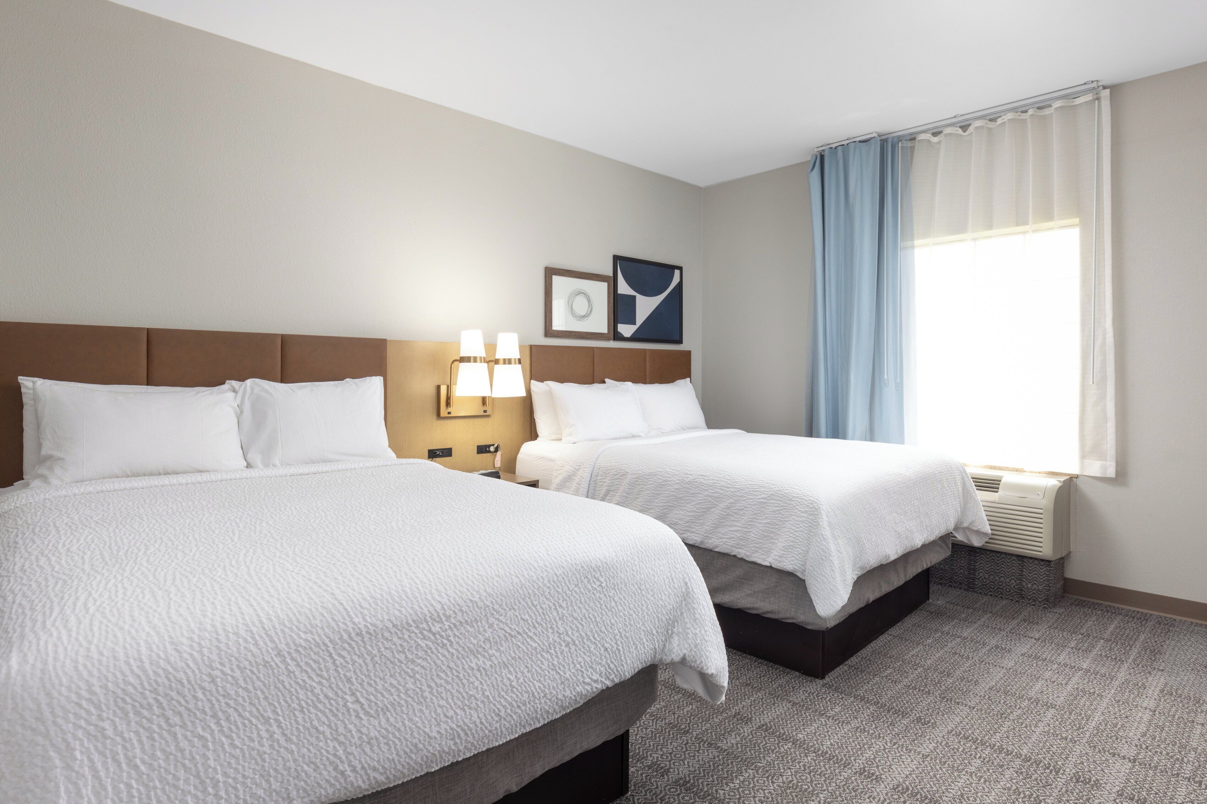 Une chambre d’hôtel avec deux lits, une fenêtre avec des rideaux, une lampe fixée au mur et un tableau encadré.