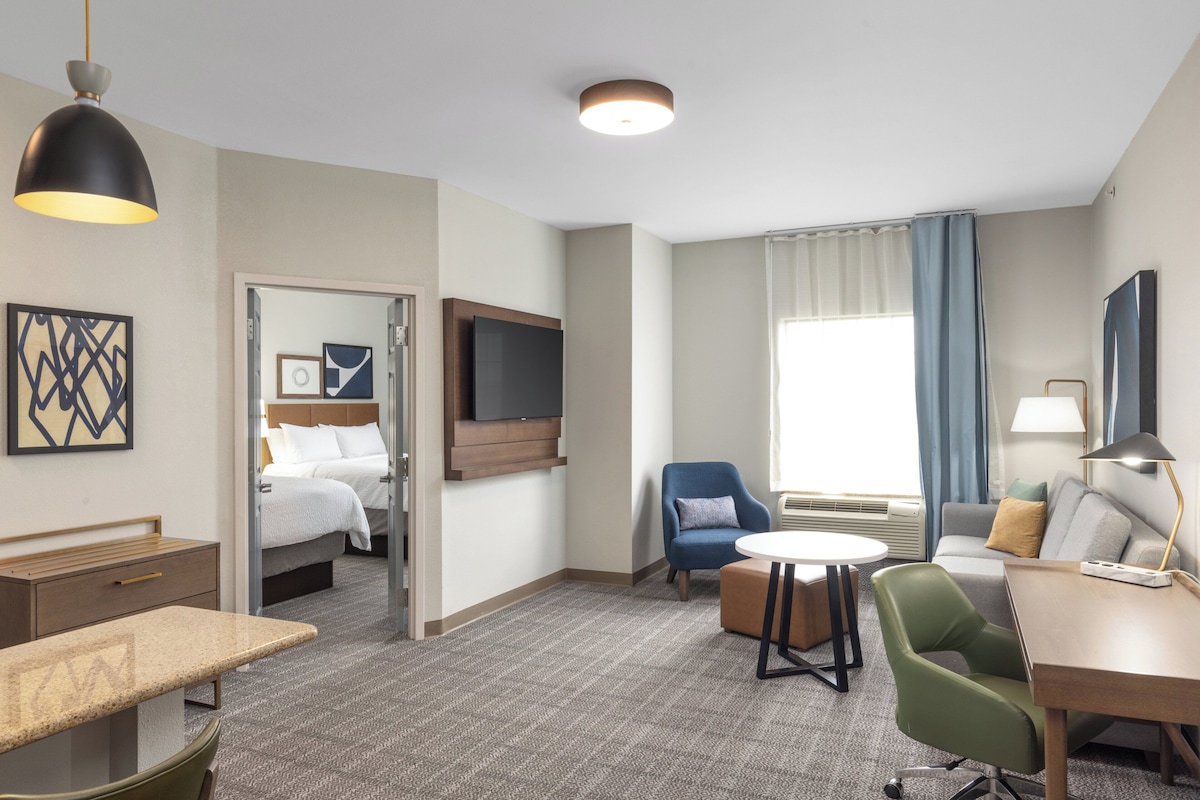 Habitación de hotel con cama, escritorio, silla, televisor y ventana con cortinas.