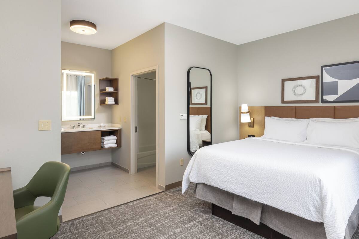 Una habitación de hotel con una cama, una silla verde, un baño con espejo y ducha.