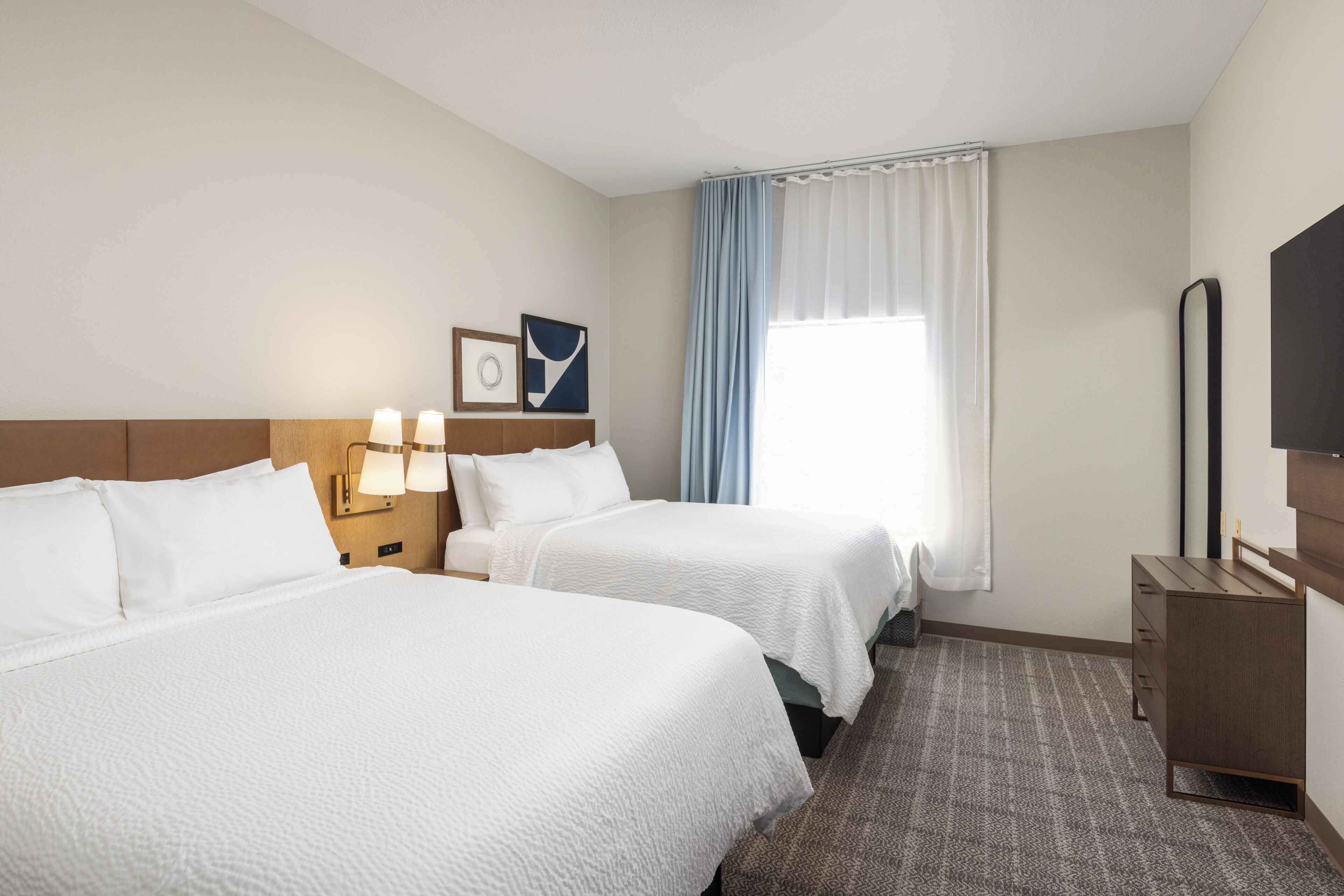 Ein Hotelzimmer mit zwei Betten, einem Nachttisch mit Lampen, einem Schminktisch, einem Spiegel und einem Fenster mit Vorhängen.