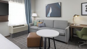 Een moderne hotelkamer met een grijze bank, een ronde witte tafel, een groene stoel, een lamp en een wanddecoratie met een abstract schilderij.