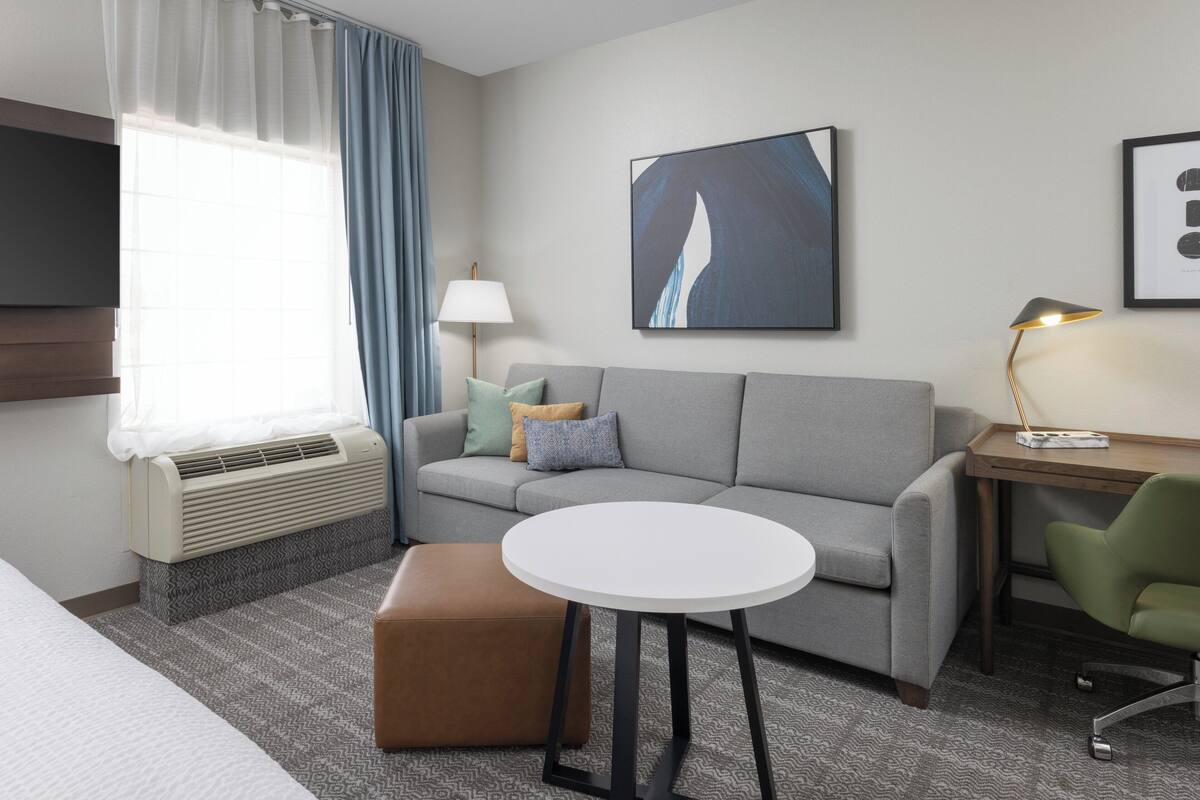 Una habitación de hotel moderna con un sofá gris, una mesa redonda blanca, una silla verde, una lámpara y un cuadro abstracto enmarcado en la pared.