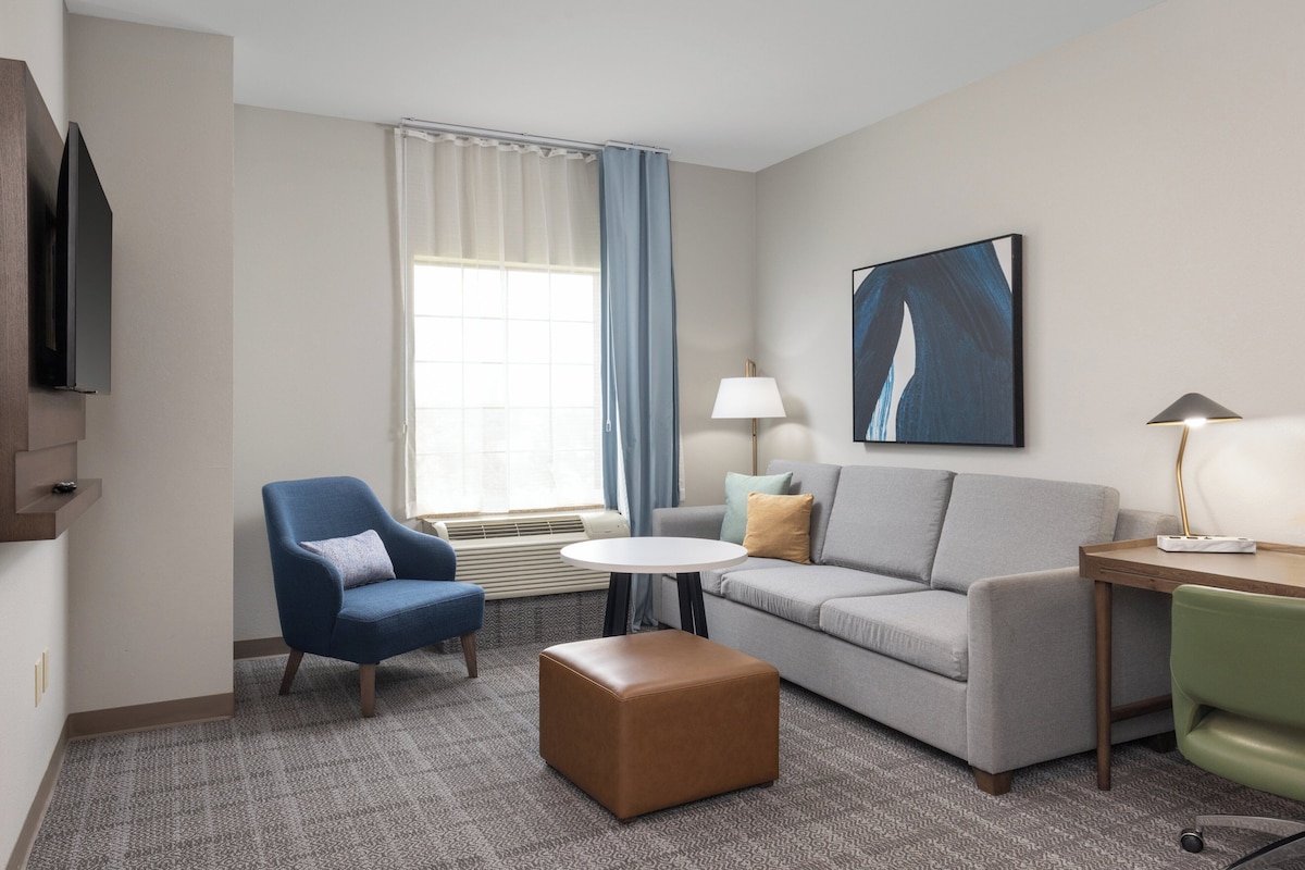 Habitación de hotel con un sofá gris, un sillón azul, un sillón verde, un reposapiés marrón, una mesita redonda pequeña, una lámpara, un cuadro, un televisor y una ventana con cortinas.
