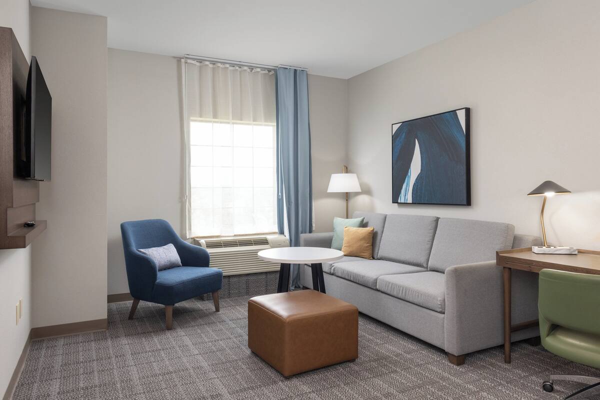 Habitación de hotel con un sofá gris, un sillón azul, un sillón verde, un reposapiés marrón, una mesita redonda pequeña, una lámpara, un cuadro, un televisor y una ventana con cortinas.