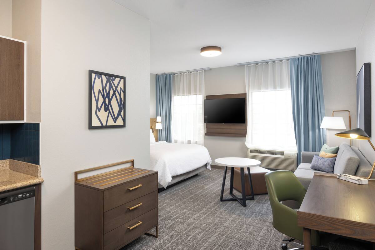 Habitación de hotel con cama, escritorio, silla, televisión y ventana con cortinas.