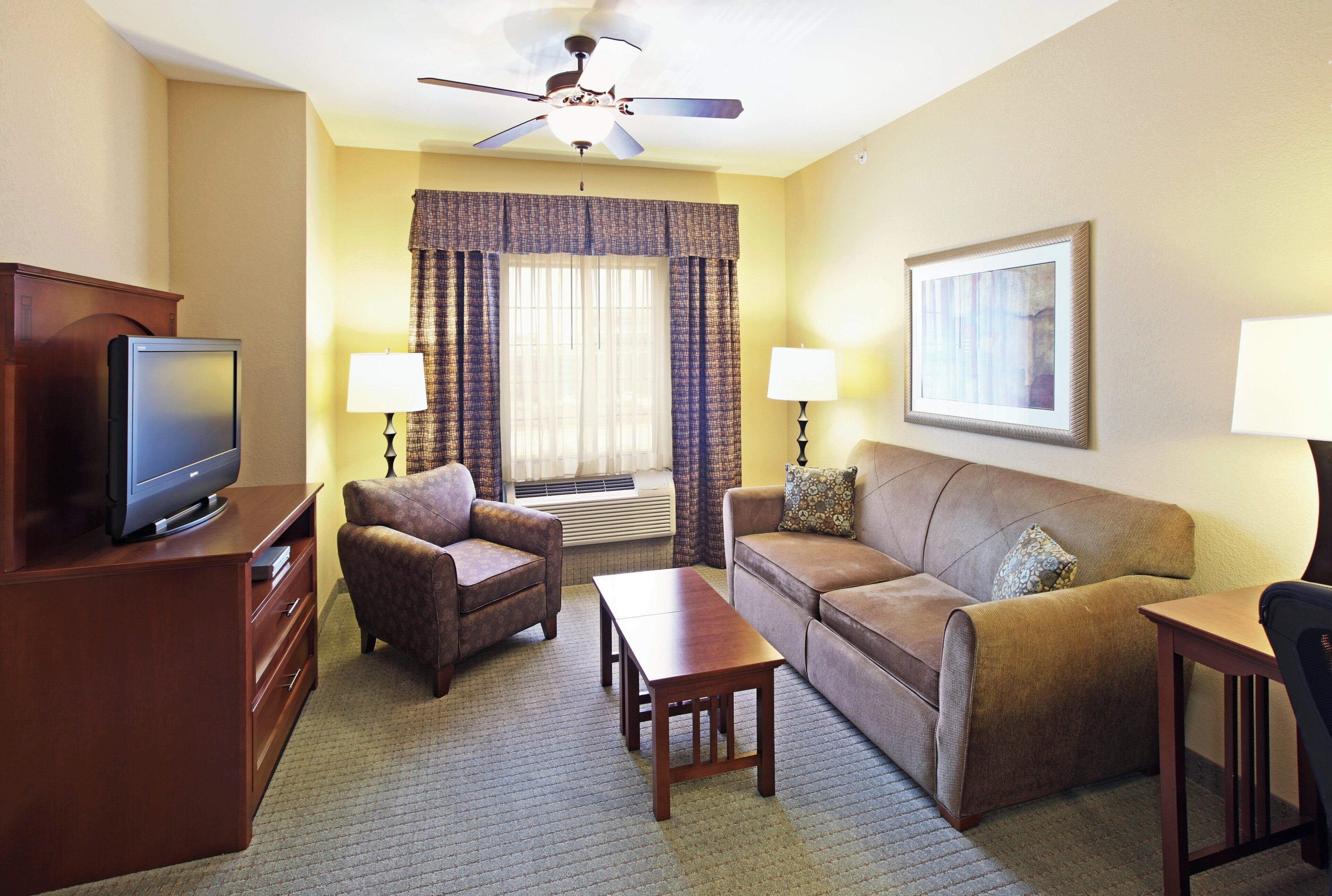 A hotel room with a flat-screen TV, a sofa, a chair, a coffee table, a ceiling fan, and a framed picture on the wall.