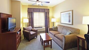 A hotel room with a flat-screen TV, a sofa, a chair, a coffee table, a ceiling fan, and a framed picture on the wall.