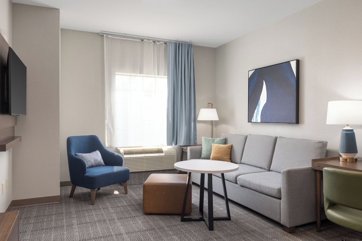 Una habitación de hotel moderna con un sofá gris, un sillón azul y una pequeña mesa redonda. Hay un televisor de pantalla plana montado en la pared, una ventana con cortinas azules y un cuadro sobre el sofá.