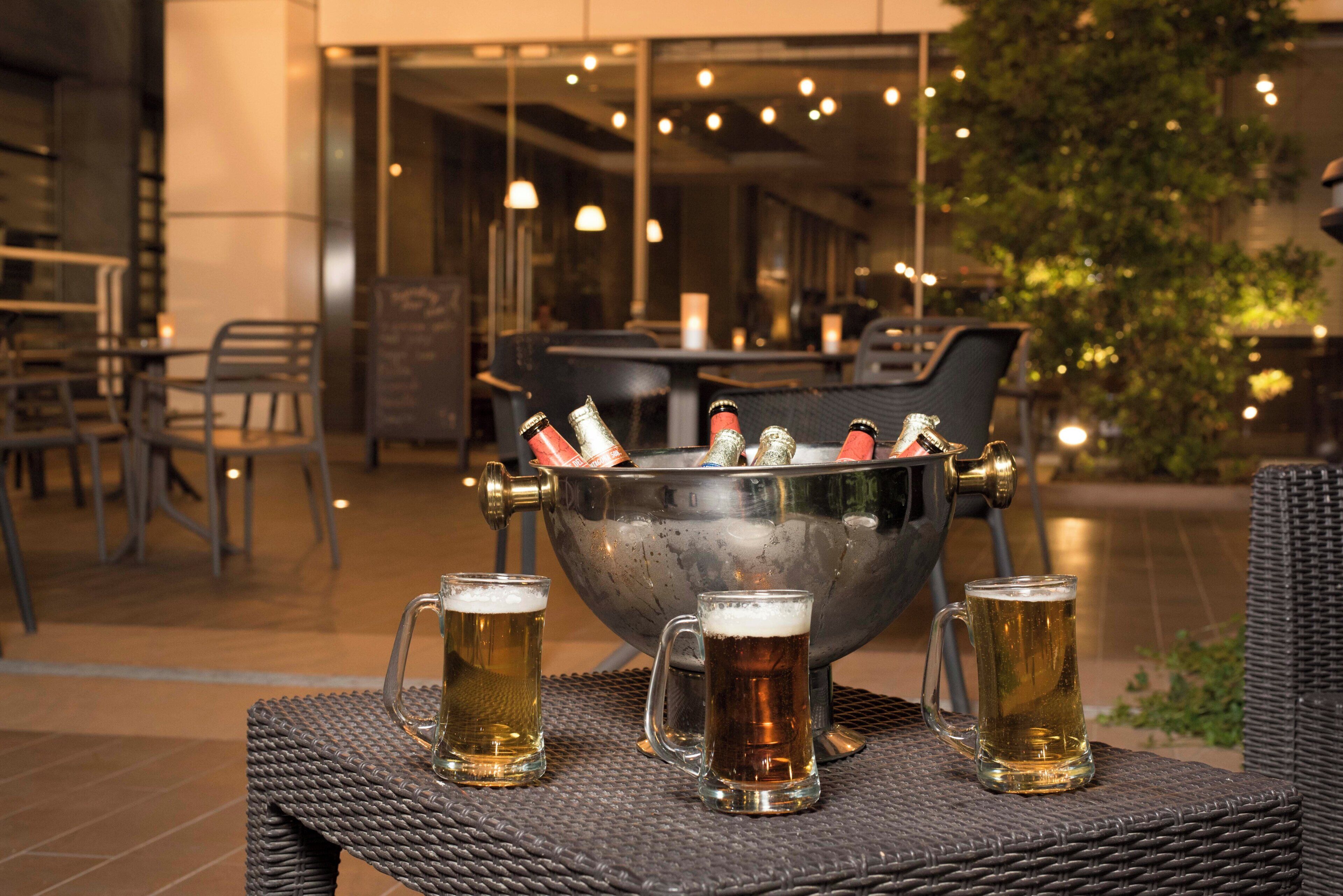 A bar area with a large metal bucket holding bottles, three glasses of beer on a wicker table, and a modern interior with chairs and tables.