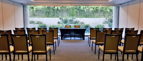 Un salón de conferencias con filas de sillas orientadas hacia una mesa, ventanas amplias con vistas a zonas verdes e iluminación empotrada en el techo.