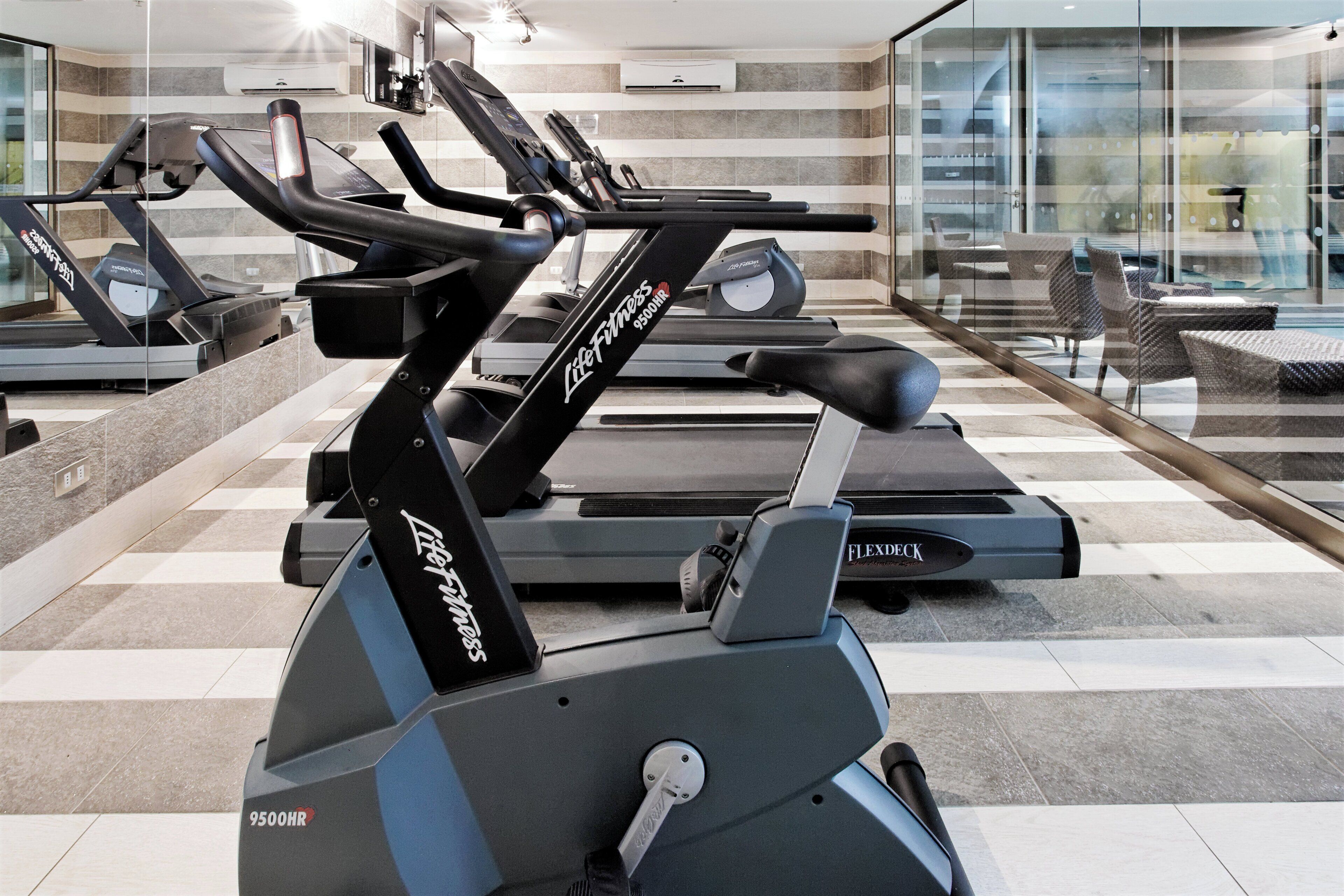 A gym with multiple treadmills and exercise bikes, a checkered floor, and a glass wall with a view of outdoor seating.