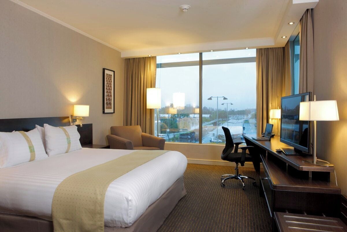 A hotel room with a large bed, a desk with a computer, a television, and a view of a cityscape through large windows.