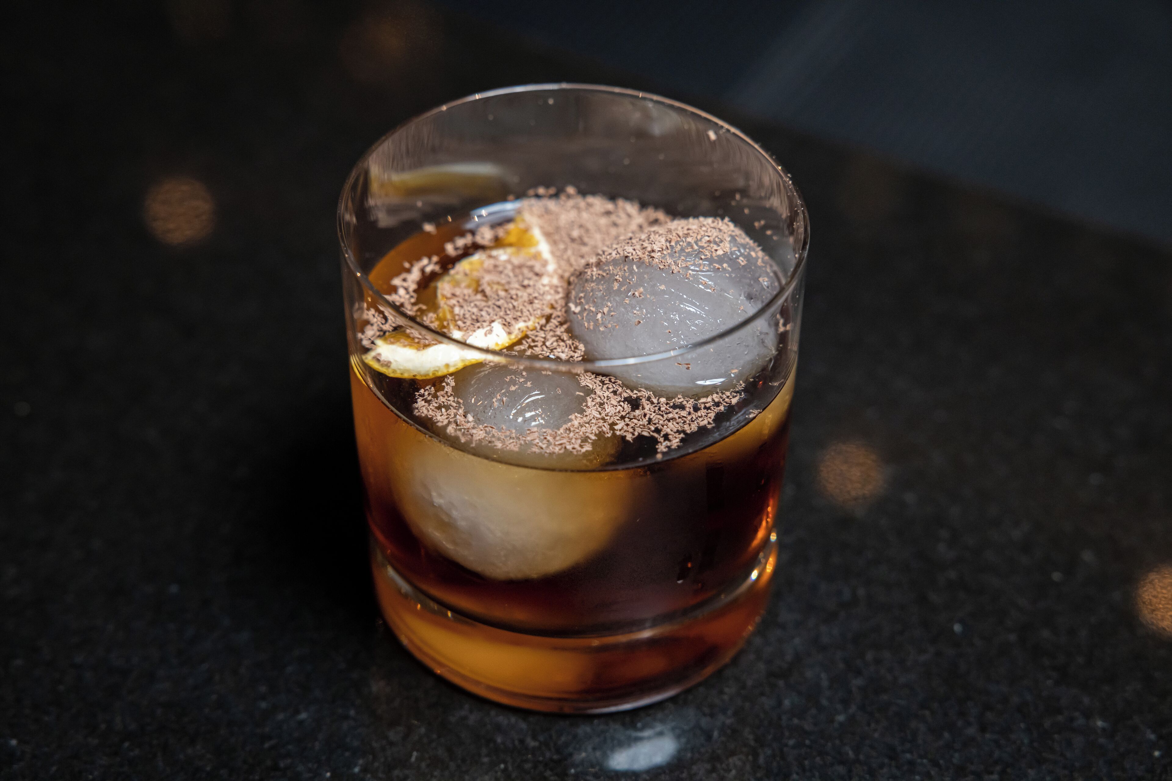 Un verre de thé glacé, une tranche de citron et une pincée de poudre brune sur une surface sombre.
