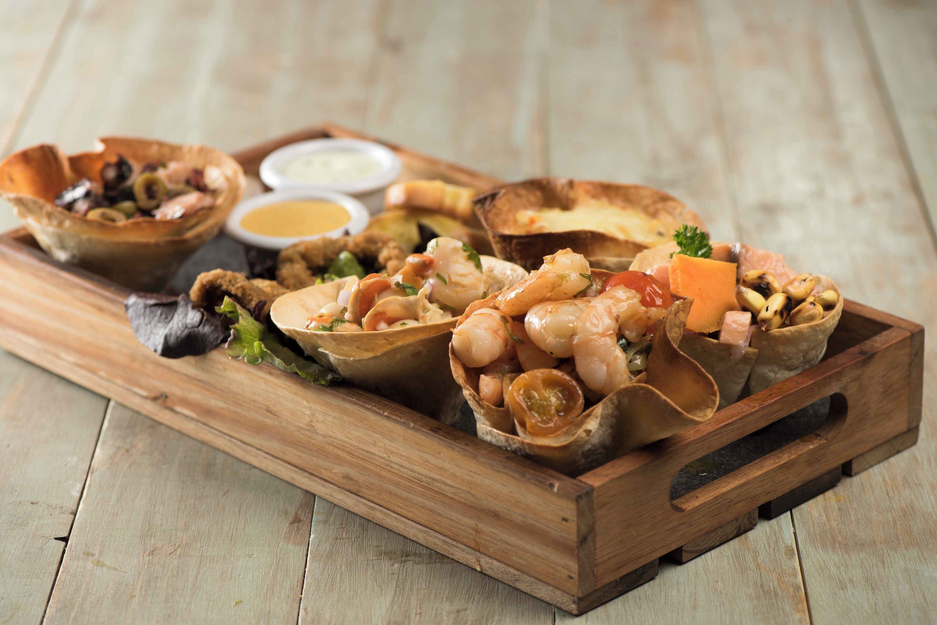 Una bandeja de madera con diversos platos de mariscos, incluyendo camarones y mejillones, acompañada de recipientes pequeños con salsas.