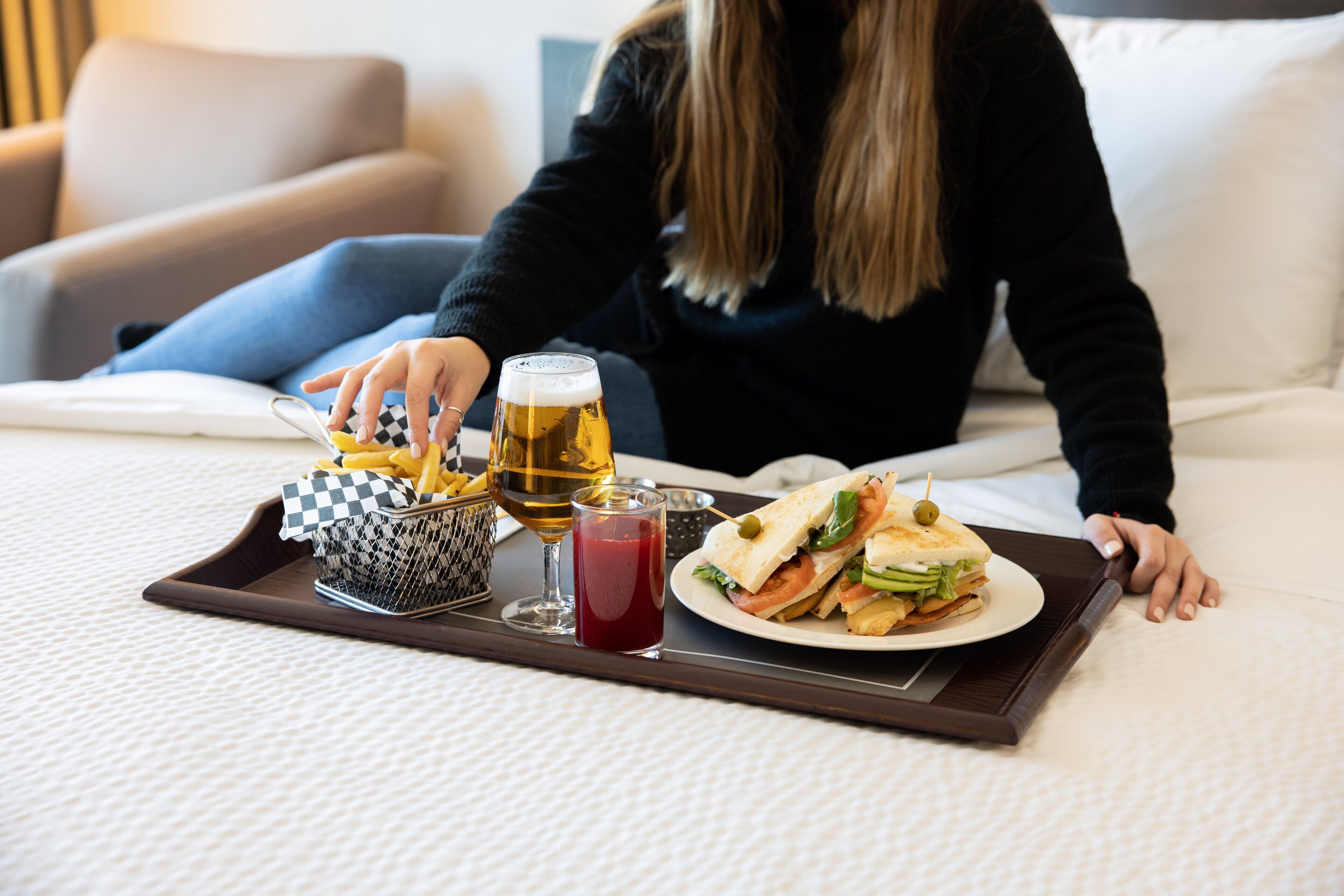A tray with a sandwich, a glass of beer, and a glass of red drink on a bed.
