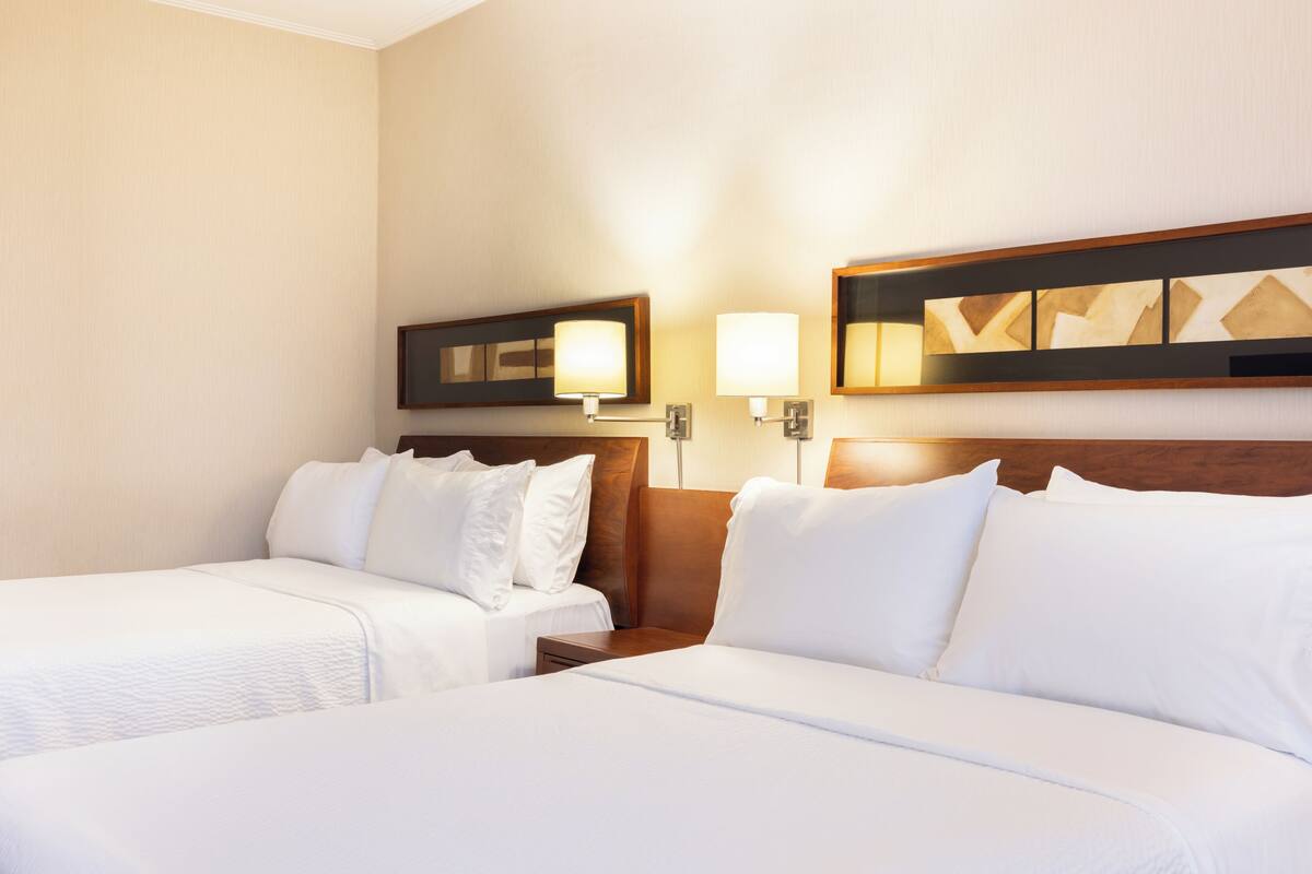 A hotel room with two beds, a wooden headboard, a nightstand, and a wall-mounted lamp.